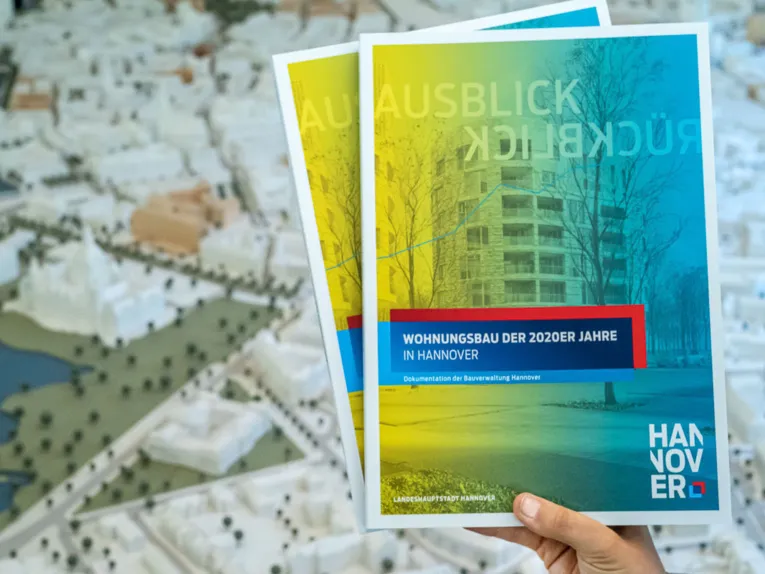 Eine Hand hält zwei Exemplare der Broschüre, im Hintergrund ein Stadtmodell